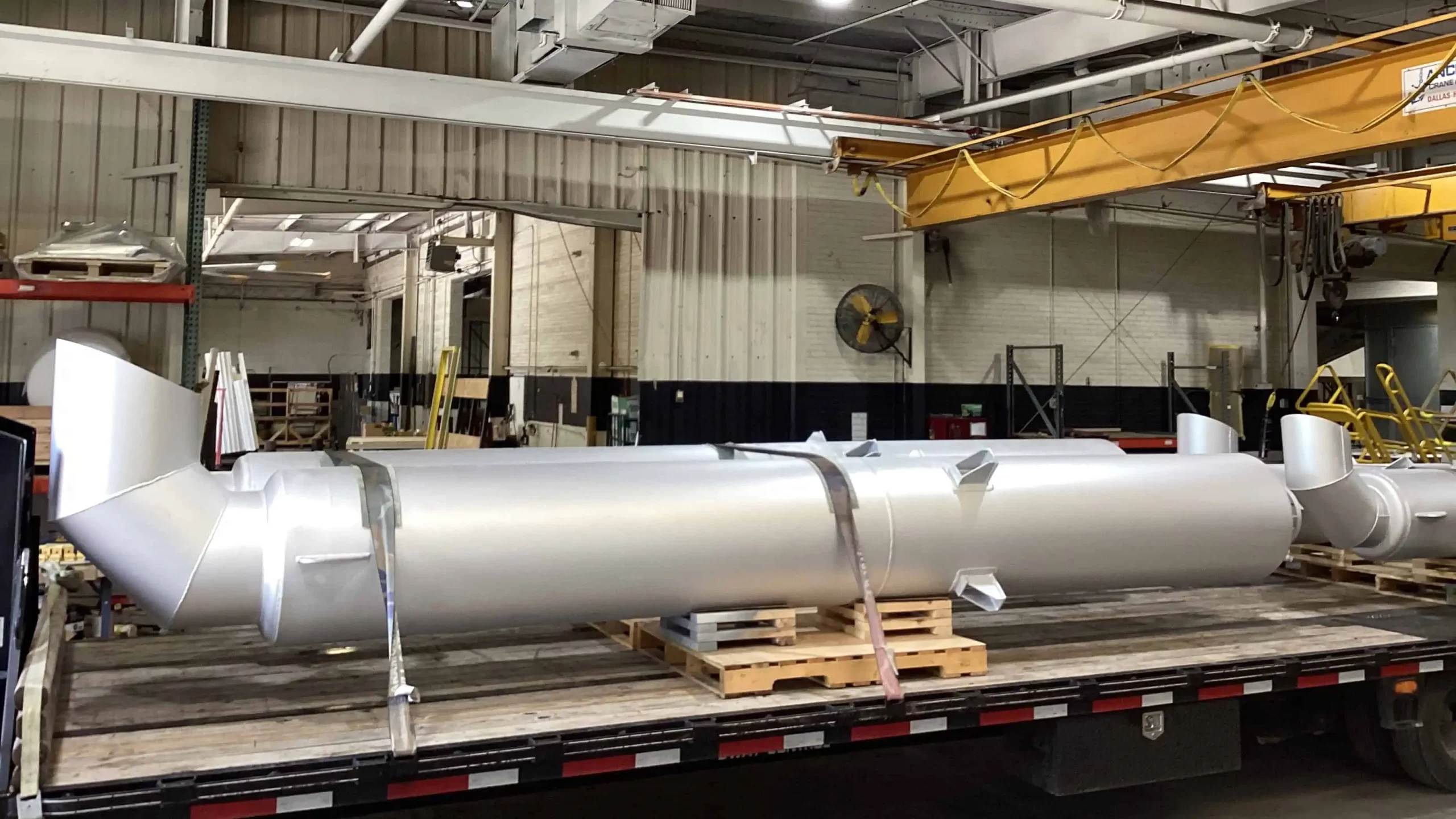 Large, freshly coated silver industrial pipes secured on a flatbed trailer inside a coating facility, with overhead cranes and various equipment in the background.