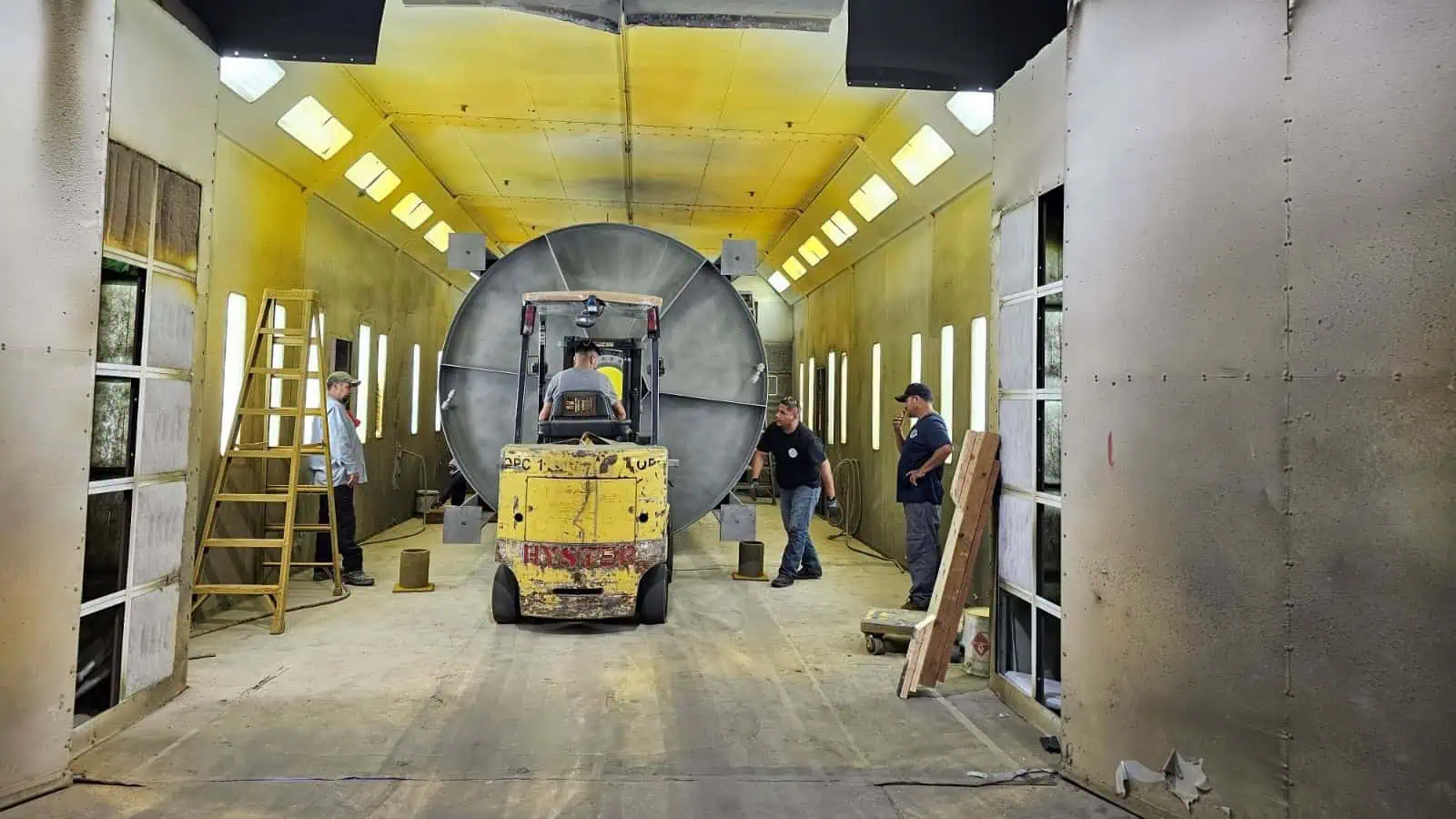 Workers in an industrial facility performing a powder coating process, with one worker operating a forklift and others overseeing the operation inside a large, well-lit coating booth.