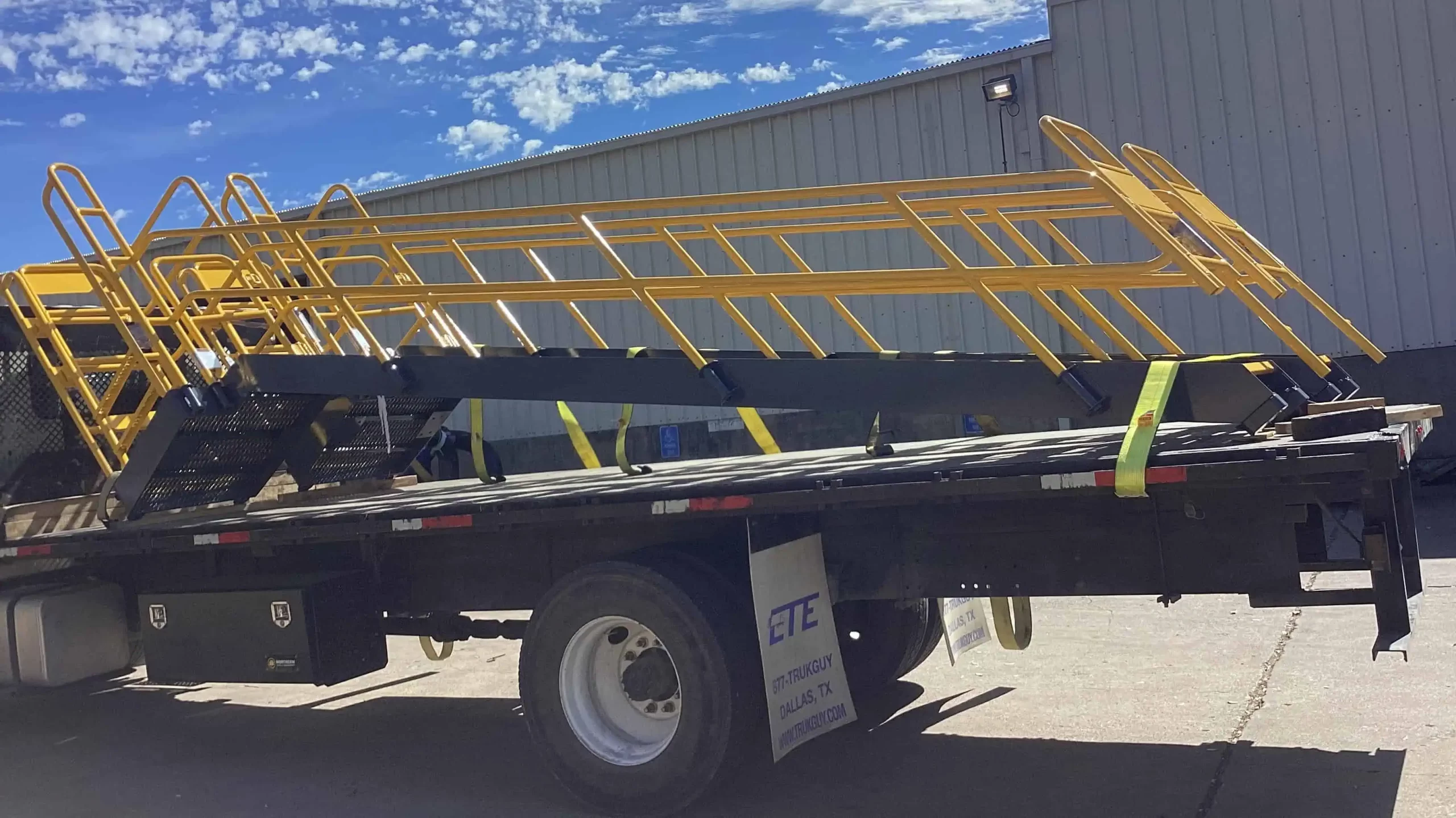 Freshly coated yellow metal stairs secured on a flatbed trailer, ready for transport, with a clear blue sky and an industrial building in the background.